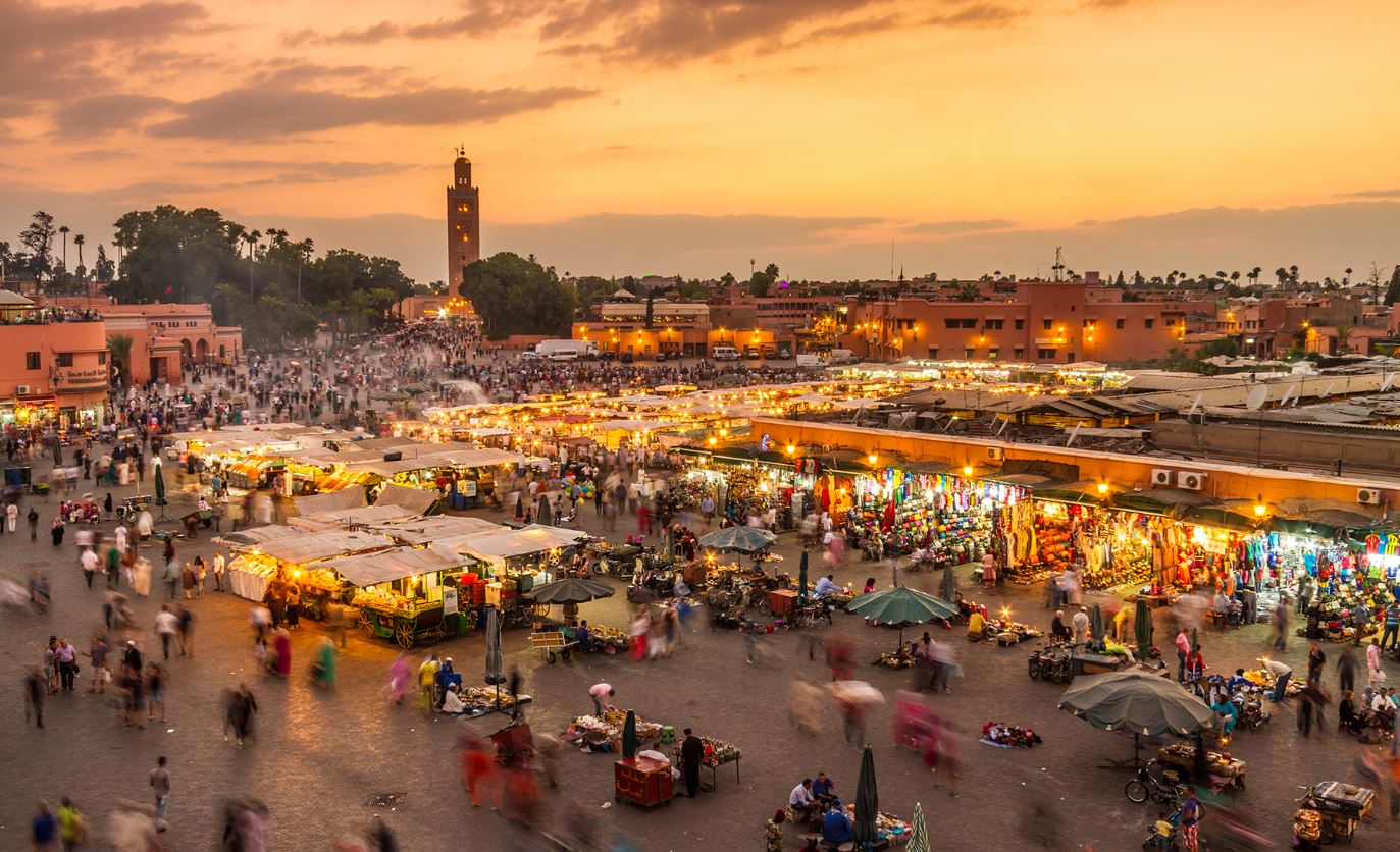 Vibrant street view of Marrakech city, Morocco, showcasing the historic Koutoubia Mosque, bustling souks, and traditional Moroccan architecture against a backdrop of palm trees and the Atlas Mountains.